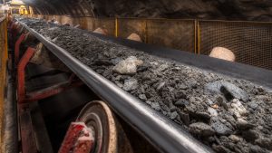 A conveyor belt in a mining tunnel transporting a mix of rocks and ore, surrounded by metal and safety barriers, with industrial rollers supporting the belt.